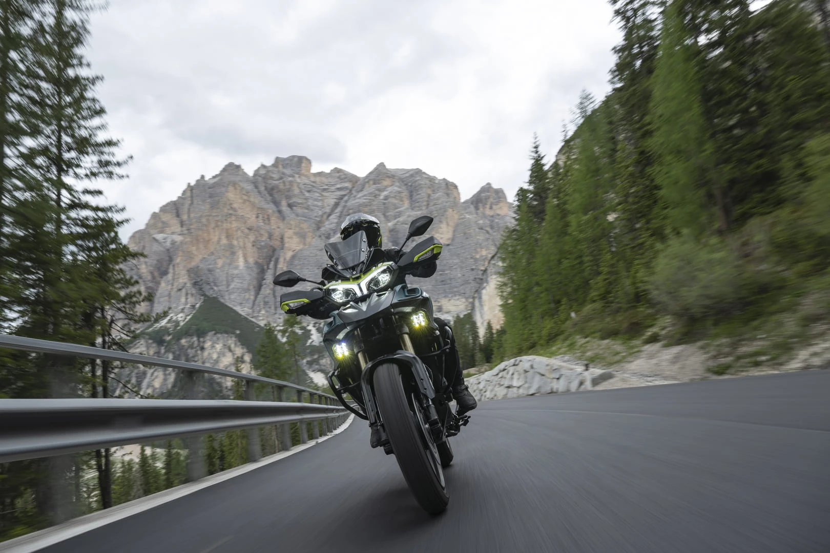 trk902xplorer_action_11 - AutoBike Motorcycle rider wearing a helmet leaning into a sharp mountain road with a guardrail and rugged cliffs in the background. - AutoBike