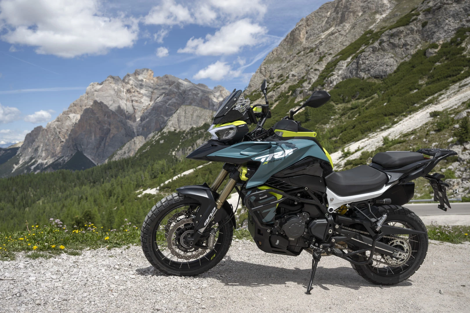 trk902xplorer_ambient_09 - AutoBike Adventure motorcycle parked on a gravel shoulder with alpine mountains and blue sky in the background. teal and neon-yellow accents on the bike show off-road styling. - AutoBike