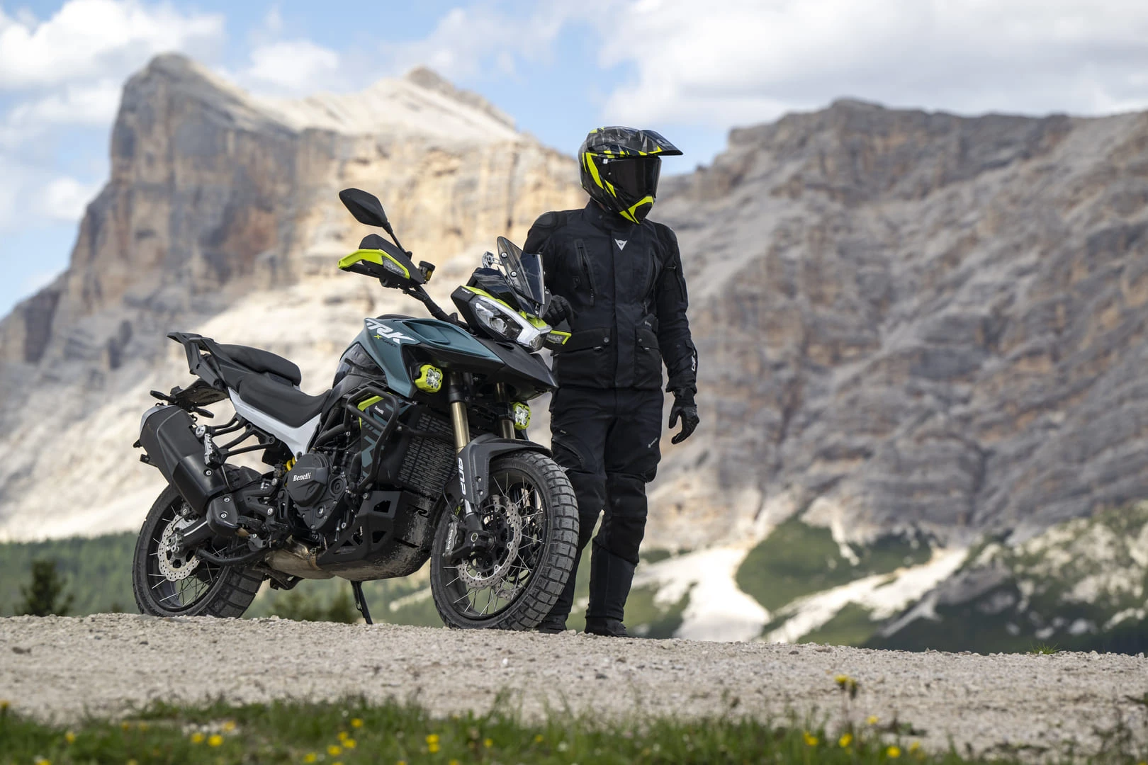 trk902xplorer_ambient_14 - AutoBike Motorcyclist in black protective gear and neon helmet beside a blue-green adventure bike on a rocky mountain landscape. - AutoBike
