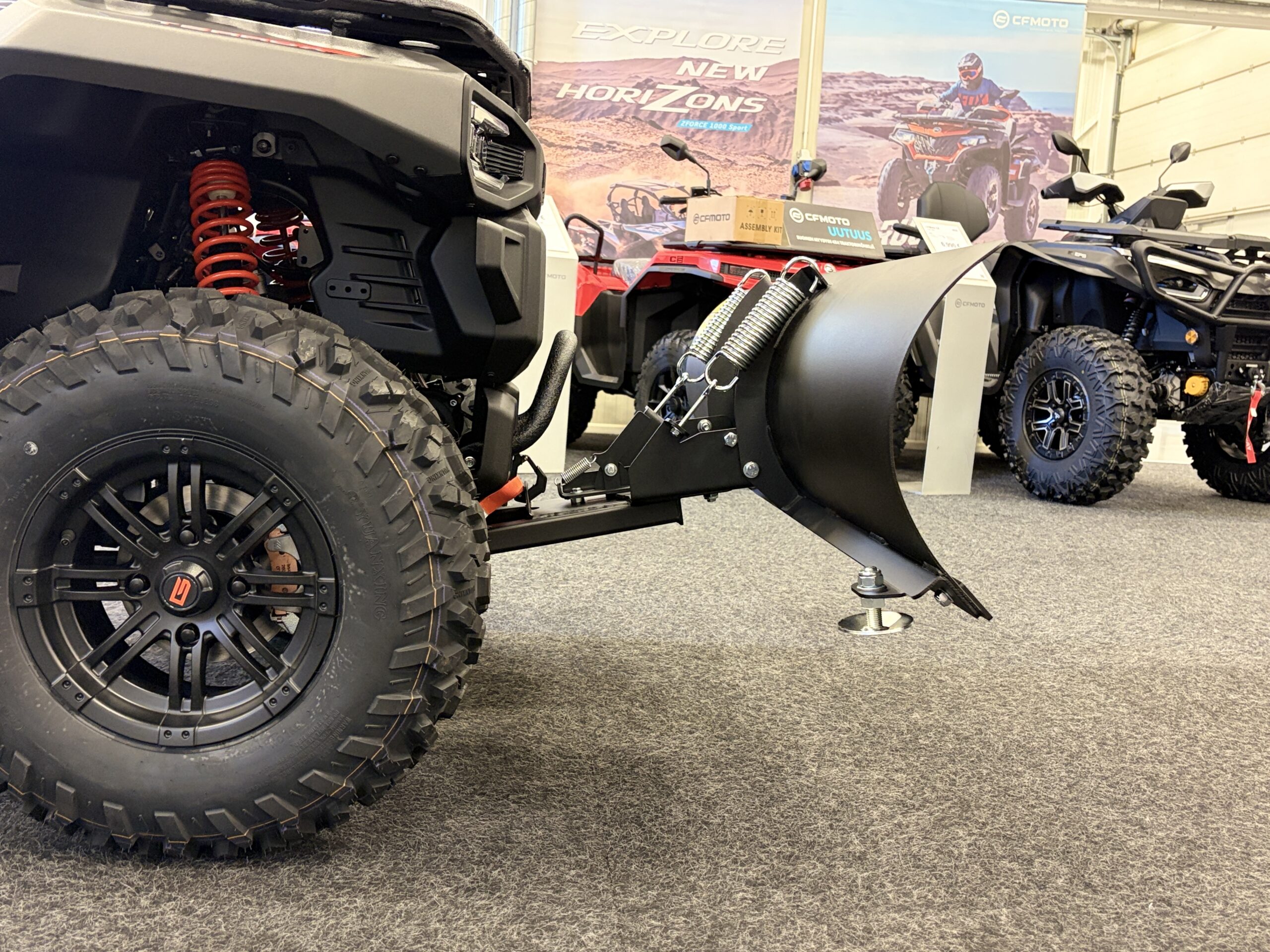 IMG_0474 - AutoBike Close-up of a rugged off-road vehicle's large treaded tire and red coil suspension in a showroom setting. - AutoBike